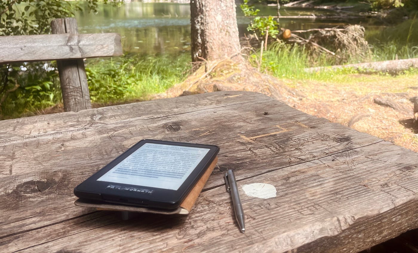 An essential kit for digital detox in nature: an e-reader, a journal, and a pen, placed on a wooden surface surrounded by natural elements.